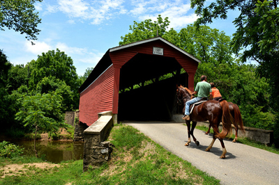 Two people riding horses on a trail in Fair Hill through a red covered bridge.